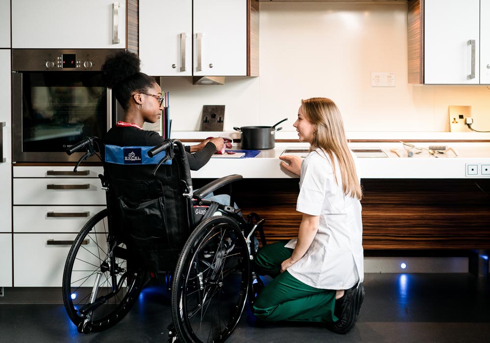 A student supporting an individual in a wheelchair in a kitchen